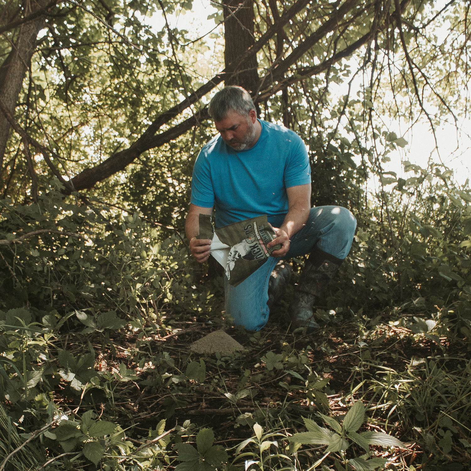 Wider photo of a man in a teal shirt pouring a bag of Black Ops Deer Anthem Molasses Granular Attractant onto the ground