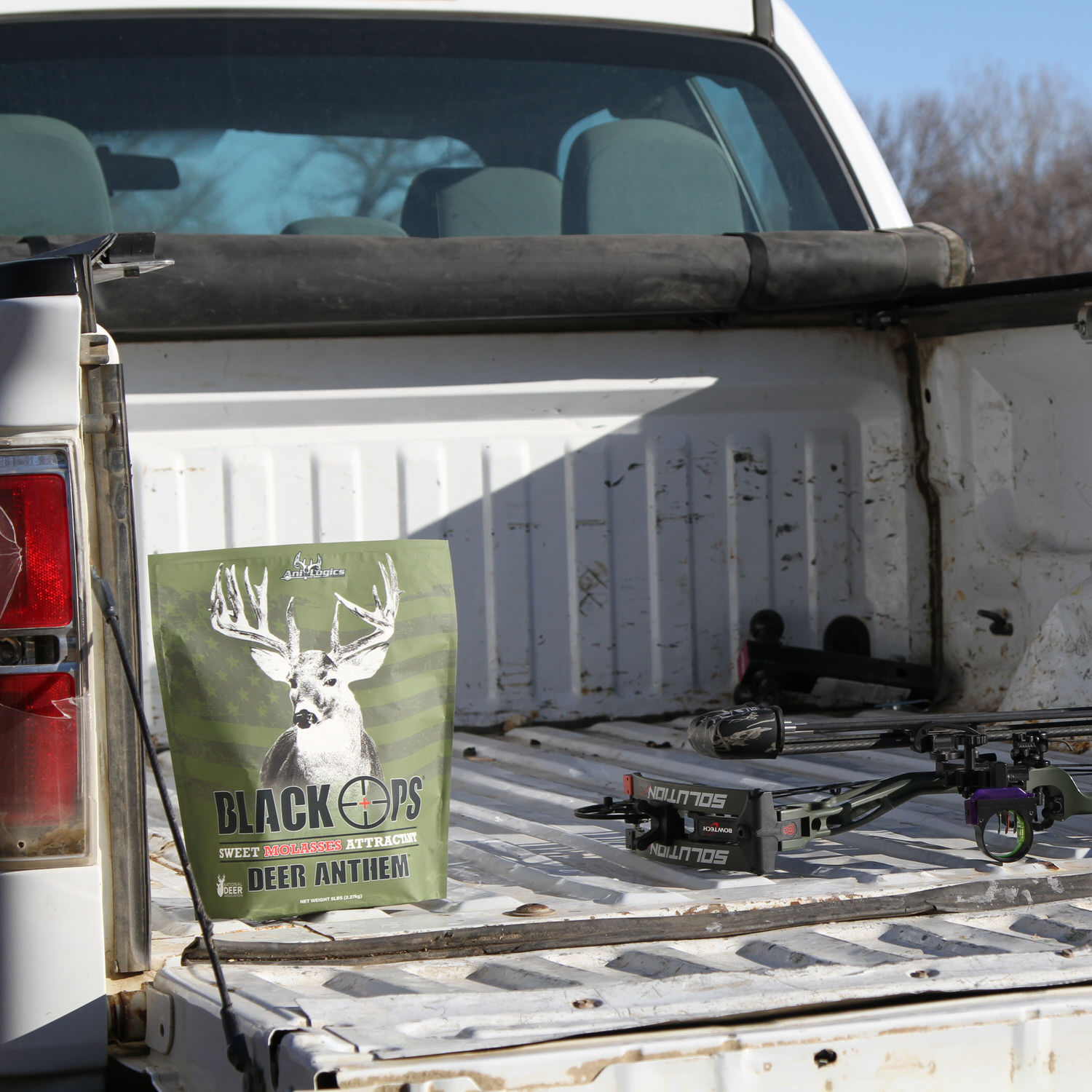 Bag of Black Ops Deer Anthem Molasses Granular Attractant in a truck bed next to a bow